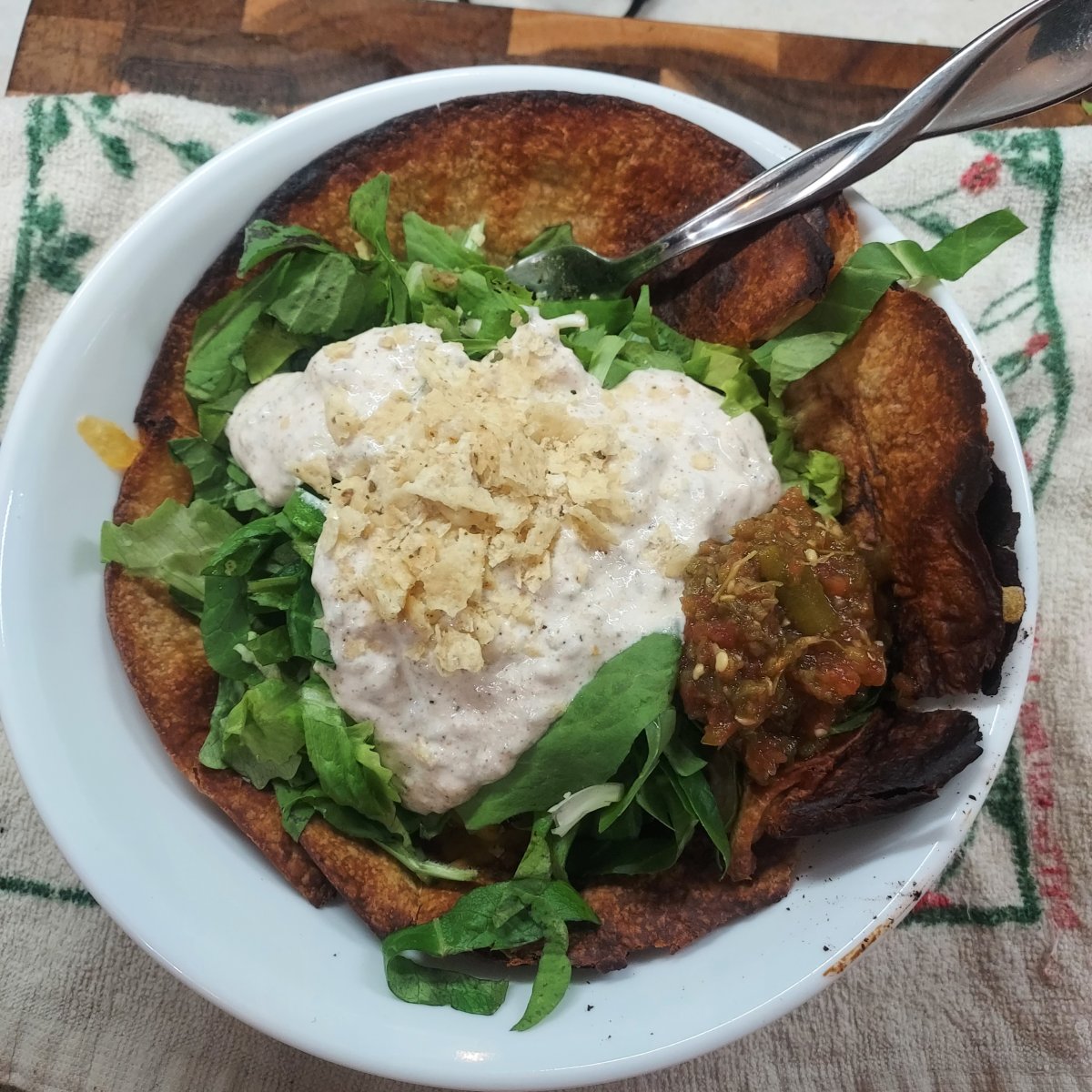 Large white bowl in the center of the photo, sitting on a worn white and green kitchen towel with a small sliver of wooden cutting board just visible beneath the towel near the top of the photo. In the bowl, a well-browned flour tortilla holds a pile of chopped raw leafy greens topped with a generous amount of white, sour cream-based salad dressing flecked with spices. The dressing is topped with a small amount of crushed white corn tortilla chips, while just to the right of the dressing and on the leafy greens is a small pile of red and green salsa.